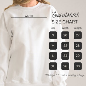 May include: A white sweatshirt with a size chart. The chart shows measurements for sizes S, M, L, and XL, with widths ranging from 20 to 26 inches and lengths from 27 to 30 inches. The text also states the model is 5'5" and wearing a large.