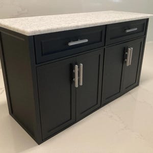 May include: A miniature kitchen cabinet unit featuring a matte black finish with two drawers and two cabinet doors. The unit is topped with a white speckled countertop and includes silver-toned bar-style handles on each drawer and door front.