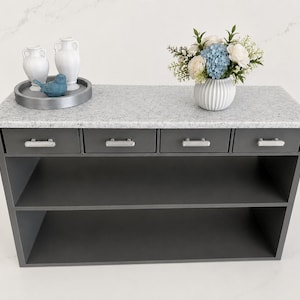 Pu&ograve; includere: Una console grigio scuro con un piano in pietra grigio chiaro maculato, quattro cassetti con maniglie argentate e due ripiani aperti. Sopra ci sono due vasi bianchi, una statuetta di uccellino blu su un vassoio grigio e un vaso con fiori.
