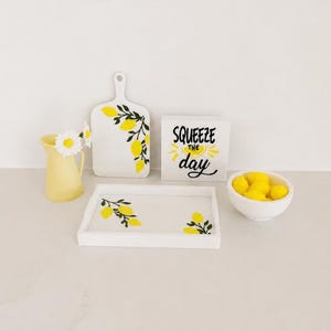 Peut inclure: Un plateau de service en bois blanc, une planche à découper blanche et un panneau carré blanc avec le texte noir SQUEEZE THE day, accompagnés d'un pichet jaune avec des fleurs blanches et un bol blanc rempli d'objets jaunes en forme de citron.