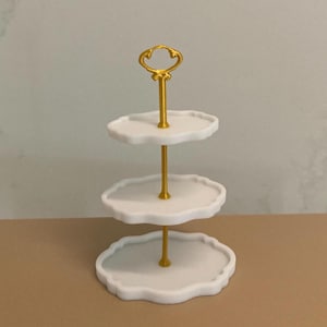 May include: A three-tiered serving tray with a gold-colored handle and support. The three white trays have a wavy edge. The serving tray is designed to hold small items such as desserts or jewelry. The tray is on a neutral-colored surface.