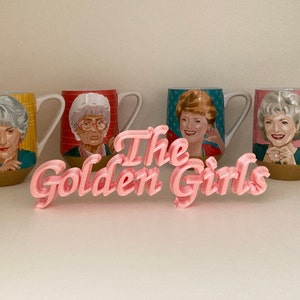 May include: Four ceramic mugs featuring portraits of four different women, each with a unique background pattern of yellow plaid, red stripes, blue stars, and pink dots. In front of the mugs sits a pink 3D-printed sign that reads The Golden Girls.