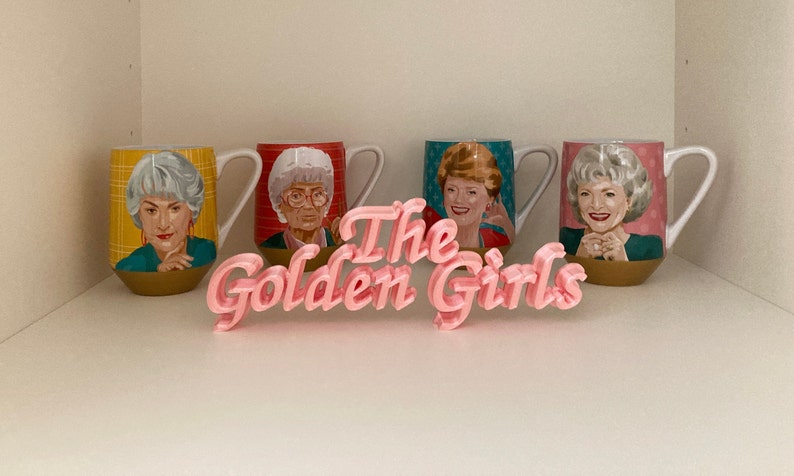 May include: Four ceramic mugs featuring portraits of four different women, each with a unique background pattern of yellow plaid, red stripes, blue stars, and pink dots. In front of the mugs sits a pink 3D-printed sign that reads The Golden Girls.