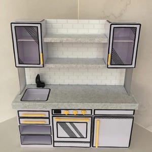 May include: A miniature toy kitchen set featuring white cabinets with black outlines, yellow handles, and a speckled gray countertop. The unit includes a sink, an oven with three yellow knobs, open shelving, and a white brick-patterned backsplash.