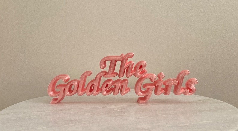 May include: A three-dimensional sign in a light pink, metallic finish featuring the text The Golden Girls in a cursive, script-style font. The letters are connected and stand upright, displaying a smooth, glossy surface with subtle horizontal ridges.