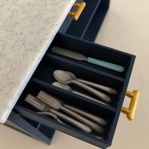 Pu&ograve; includere: Un cassetto da cucina in miniatura blu scuro con piano di lavoro bianco maculato e maniglie color oro. Il cassetto aperto &egrave; diviso in scomparti contenenti piccole posate color argento, tra cui forchette, cucchiai e un coltello con manico.