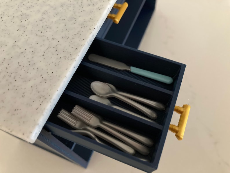 Pu&ograve; includere: Un cassetto da cucina in miniatura blu scuro con piano di lavoro bianco maculato e maniglie color oro. Il cassetto aperto &egrave; diviso in scomparti contenenti piccole posate color argento, tra cui forchette, cucchiai e un coltello con manico.