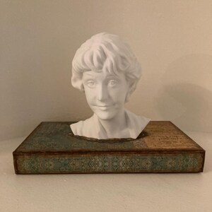 May include: A white bust of a woman with curly hair sits atop a decorative box with a teal and gold design. The bust is a classical sculpture, and the box has an aged, vintage appearance.