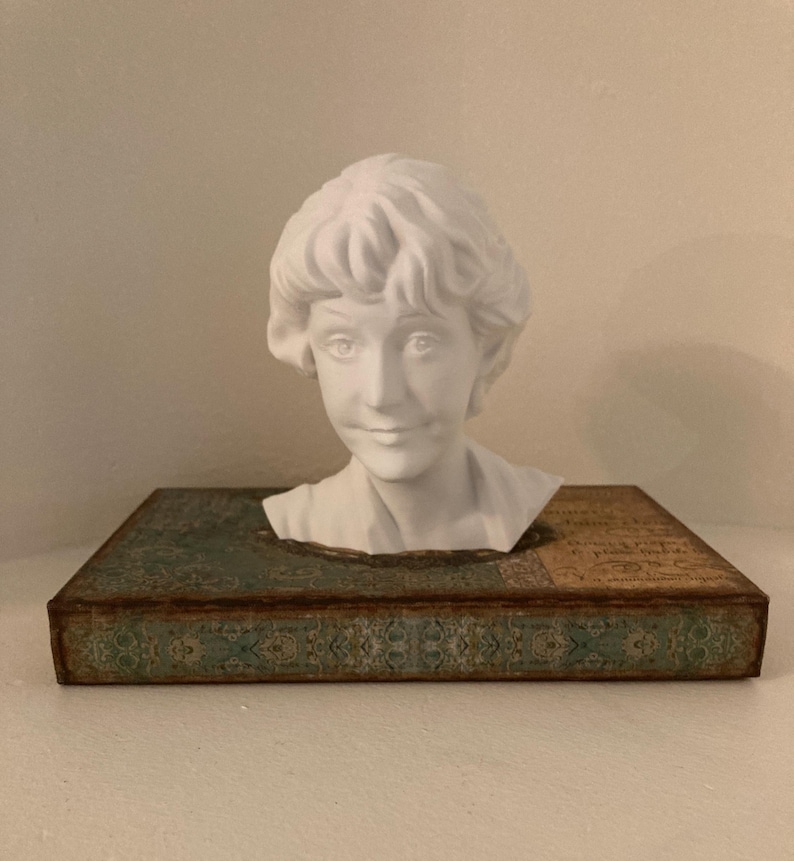 May include: A white bust of a woman with curly hair sits atop a decorative box with a teal and gold design. The bust is a classical sculpture, and the box has an aged, vintage appearance.
