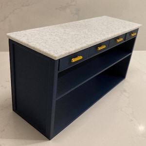 Pu&ograve; includere: Un'isola da cucina in miniatura con una base blu navy scuro e tre piccoli cassetti dotati di maniglie orizzontali color oro. La superficie superiore &egrave; una lastra rettangolare piatta con una texture maculata bianca e grigia.