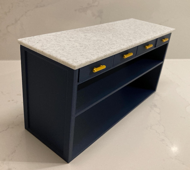 Pu&ograve; includere: Un'isola da cucina in miniatura con una base blu navy scuro e tre piccoli cassetti dotati di maniglie orizzontali color oro. La superficie superiore &egrave; una lastra rettangolare piatta con una texture maculata bianca e grigia.