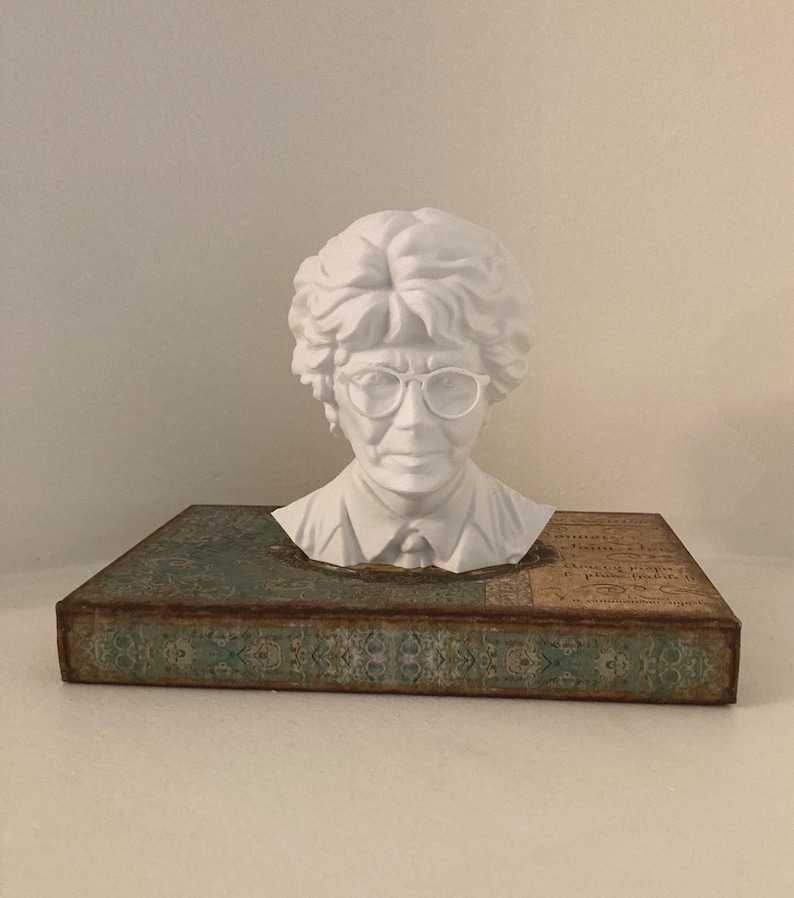 May include: A white bust of a person wearing glasses, atop a decorative book. The bust has detailed facial features and curly hair. The book has a teal and brown patterned cover with aged edges, adding a vintage aesthetic.