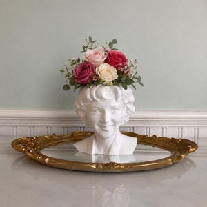 Puede incluir: Un busto de cerámica blanca de una mujer con un arreglo floral de rosas rosas y crema. El busto se encuentra sobre una bandeja ovalada dorada. Las flores son una mezcla de rosas rosas claras y oscuras y crema.