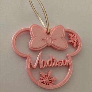 May include: Pink plastic bauble shaped like a mouse head with a bow and the name "Madison" written inside. The bauble has snowflakes cut out of it.