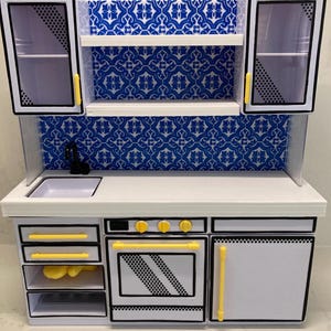 May include: A miniature dollhouse kitchen unit featuring white cabinets with black outlines, yellow handles, and a blue and white patterned backsplash. The set includes an oven, a sink with a black faucet, open shelving, and storage drawers.