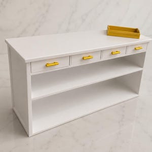 Pu&ograve; includere: Un tavolino console in miniatura bianco con quattro piccoli cassetti dotati di maniglie orizzontali dorate allineate nella parte superiore. Sotto i cassetti ci sono due ripiani aperti. Un piccolo vassoio rettangolare dorato poggia sopra.