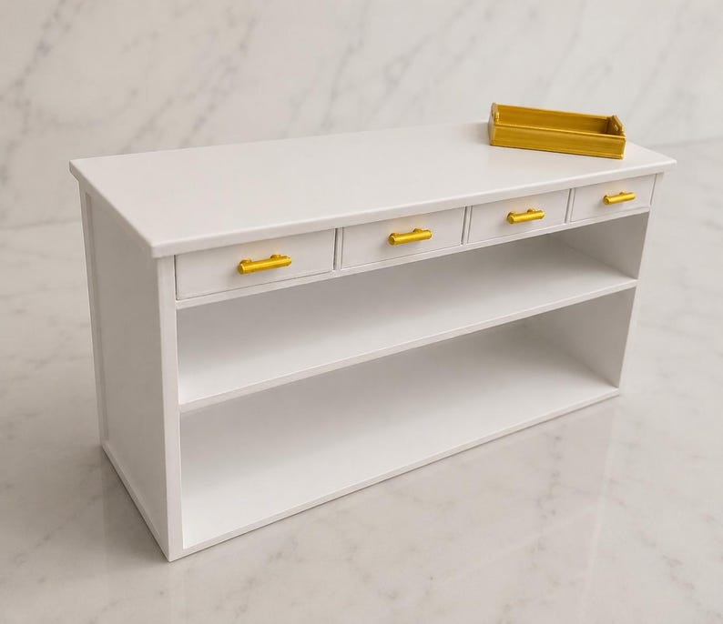 Pu&ograve; includere: Un tavolino console in miniatura bianco con quattro piccoli cassetti dotati di maniglie orizzontali dorate allineate nella parte superiore. Sotto i cassetti ci sono due ripiani aperti. Un piccolo vassoio rettangolare dorato poggia sopra.