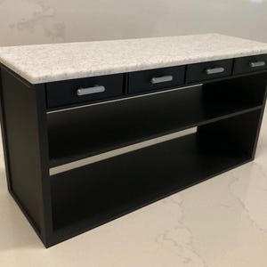 Pu&ograve; includere: Un'isola da cucina in miniatura con struttura e base nere e due ripiani aperti. La superficie superiore &egrave; un piano di lavoro in finto marmo bianco maculato. Sotto il piano si trovano quattro cassetti neri, ciascuno con una maniglia argentata.