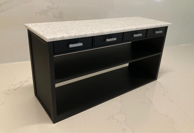 Pu&ograve; includere: Un'isola da cucina in miniatura con struttura e base nere e due ripiani aperti. La superficie superiore &egrave; un piano di lavoro in finto marmo bianco maculato. Sotto il piano si trovano quattro cassetti neri, ciascuno con una maniglia argentata.