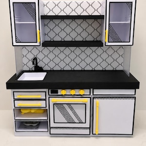 May include: A miniature toy kitchen set featuring white cabinets with black trim, yellow handles, and a black countertop. The backsplash has a gray and white quatrefoil pattern. The unit includes an oven, sink, and open shelving with black shelves.