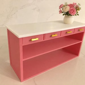 Pu&ograve; includere: Una console in miniatura rosa con un piano rettangolare bianco, tre piccoli cassetti con maniglie orizzontali dorate e due ripiani aperti. Sopra &egrave; posizionato un piccolo vaso bianco a coste contenente fiori artificiali rosa e bianchi.
