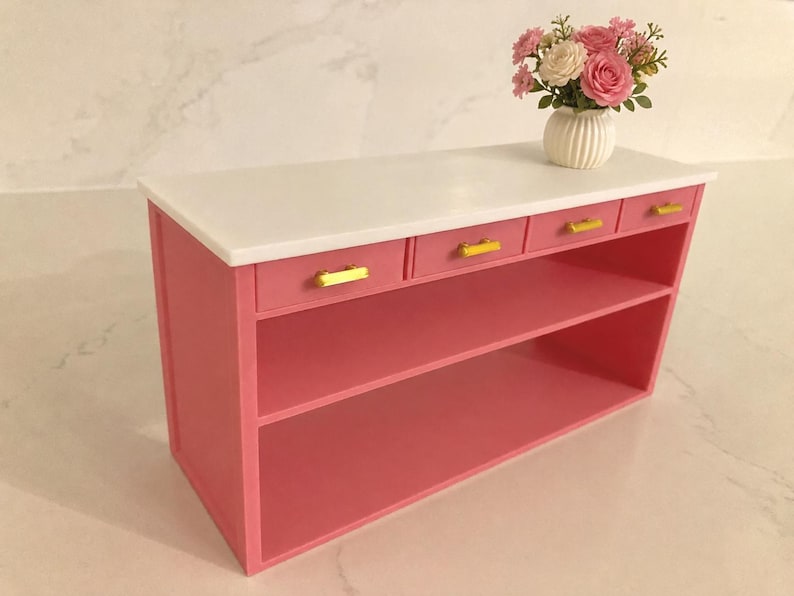 Pu&ograve; includere: Una console in miniatura rosa con un piano rettangolare bianco, tre piccoli cassetti con maniglie orizzontali dorate e due ripiani aperti. Sopra &egrave; posizionato un piccolo vaso bianco a coste contenente fiori artificiali rosa e bianchi.
