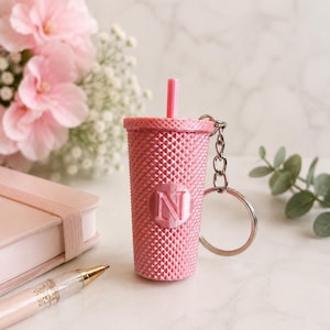 May include: A pink tumbler-shaped keyring featuring a textured, diamond-patterned surface and a small straw on top. The centre of the tumbler displays a circular emblem with the letter N embossed in the middle. A silver metal ring is attached.