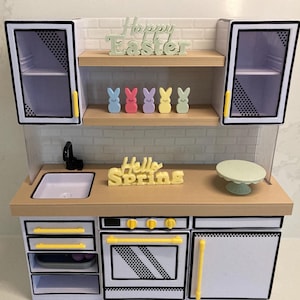 May include: A white toy kitchen set with a black and white patterned design. The kitchen has a sink, oven, and two cabinets. The top of the kitchen has a shelf with a sign that says "Happy Easter" and another shelf with a sign that says "Hello Spring".