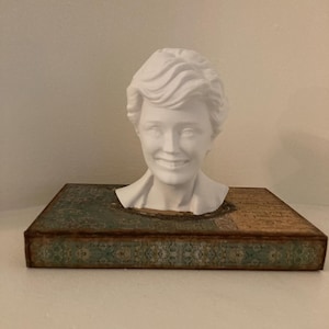 May include: A white bust of a smiling woman with short, wavy hair sits atop a decorative box. The box has a teal and brown patterned design with a distressed, vintage look. The bust is a detailed sculpture.