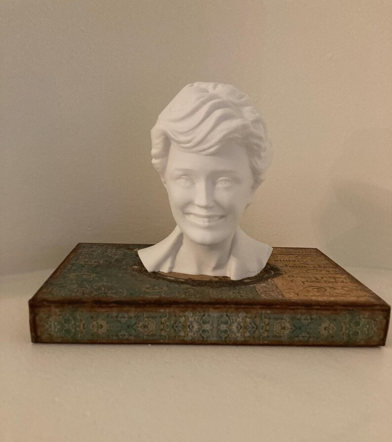 May include: A white bust of a smiling woman with short, wavy hair sits atop a decorative box. The box has a teal and brown patterned design with a distressed, vintage look. The bust is a detailed sculpture.