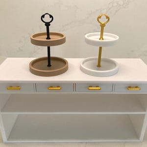 Könnte beinhalten: Zweistöckige Servierstände, einer beige und schwarz, der andere weiß und gold, stehen auf einem weißen Tisch mit goldenen Schubladengriffen. Die Ständer haben ein kreisförmiges Design, ideal für die Präsentation kleiner Gegenstände oder Leckereien.