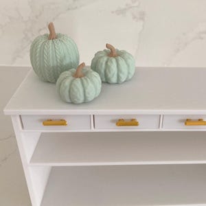 May include: Three light mint green decorative pumpkins with a textured knit-like pattern and tan stems, arranged on top of a white miniature console table featuring three drawers with gold-toned horizontal handles and an open lower shelf.