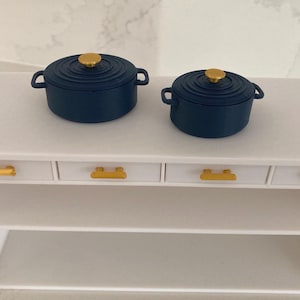 May include: Two navy blue miniature cast iron pots with gold-colored knobs and handles sit on a white table. The table has four drawers with gold-colored handles and two shelves.