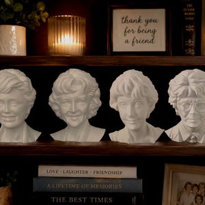 May include: Four white busts of women with different hairstyles and expressions are displayed on a dark wooden shelf. The busts are arranged in a row. A framed sign above reads "thank you for being a friend."