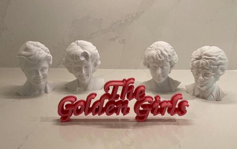 May include: Four white busts of the Golden Girls characters with the pink text "The Golden Girls" in front. The busts are of Dorothy, Rose, Blanche, and Sophia. The busts are on a white surface.