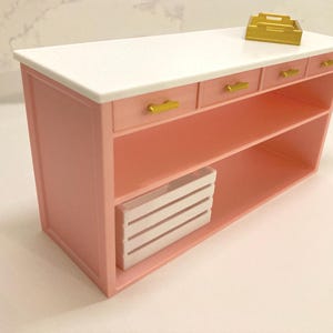 Pu&ograve; includere: Un'isola da cucina in miniatura rosa con piano di lavoro bianco, tre piccoli cassetti con maniglie dorate, un ripiano aperto e una cassetta a doghe bianca all'interno. Un piccolo vassoio dorato &egrave; posizionato sulla superficie superiore.