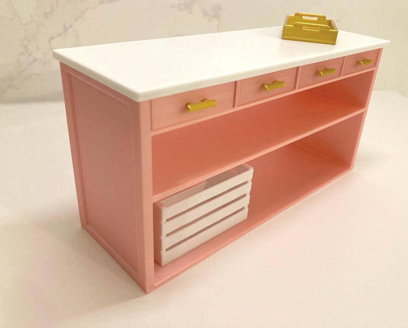 Pu&ograve; includere: Un'isola da cucina in miniatura rosa con piano di lavoro bianco, tre piccoli cassetti con maniglie dorate, un ripiano aperto e una cassetta a doghe bianca all'interno. Un piccolo vassoio dorato &egrave; posizionato sulla superficie superiore.