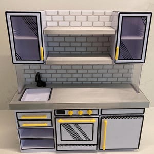 May include: A toy kitchen playset featuring a white and gray color scheme with yellow accents. The unit includes upper cabinets with black trim, open shelving, a sink with a black faucet, a stove with three yellow knobs, and lower storage drawers.