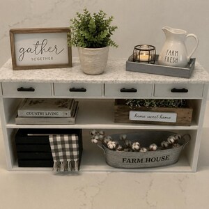 Pu&ograve; includere: Un tavolo consolle bianco con piano effetto marmo, quattro cassetti con maniglie nere e due ripiani aperti. L'arredamento include un cartello gather together, una pianta, una candela, una brocca farm fresh, libri e un contenitore in metallo.
