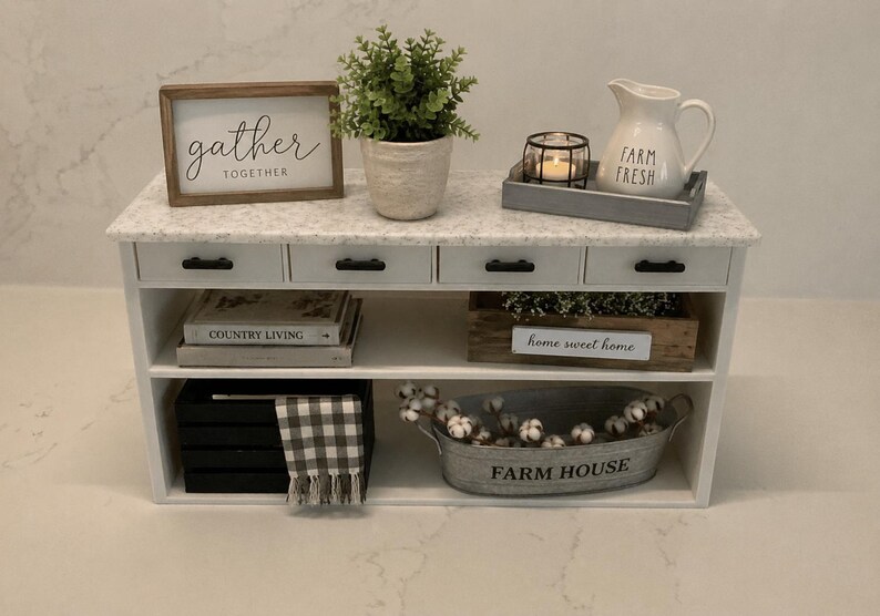 Pu&ograve; includere: Un tavolo consolle bianco con piano effetto marmo, quattro cassetti con maniglie nere e due ripiani aperti. L'arredamento include un cartello gather together, una pianta, una candela, una brocca farm fresh, libri e un contenitore in metallo.