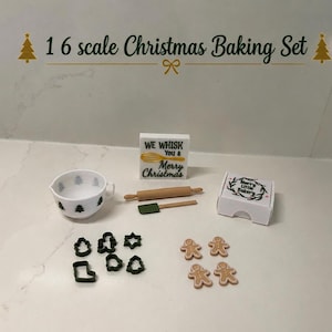 May include: A 1:6 scale Christmas baking set featuring a white mixing bowl with tree patterns, a wooden rolling pin, a green spatula, a sign reading We Whisk You a Merry Christmas, a box labelled Merry Little Bakery, six dark green cookie cutters, and four gingerbread man figures.