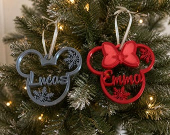 May include: Two mouse-shaped holiday ornaments with hanging ribbons. The gray ornament features the name Lucas and snowflake cutouts. The red ornament features the name Emma, a bow detail at the top, and snowflake cutouts. Both are made of plastic.