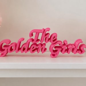 May include: A bright pink, three-dimensional sign featuring the text The Golden Girls in a stylized, cursive font. The letters are connected and stand upright, creating a bold display piece with a smooth, matte finish and distinct depth.