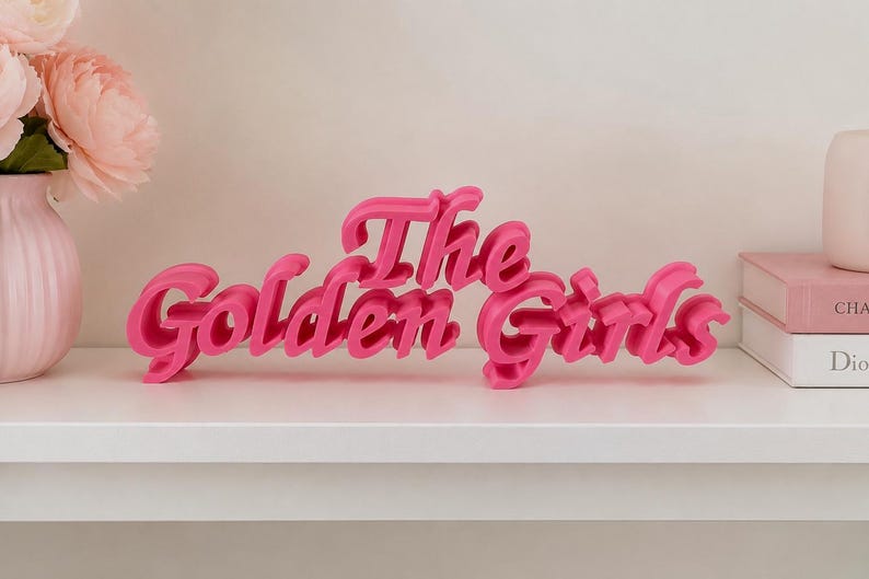 May include: A bright pink, three-dimensional sign featuring the text The Golden Girls in a stylized, cursive font. The letters are connected and stand upright, creating a bold display piece with a smooth, matte finish and distinct depth.