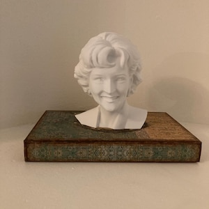 May include: A white bust of a smiling woman with curly hair rests on a rectangular box. The box has a teal and brown patterned design. The bust is a decorative item.