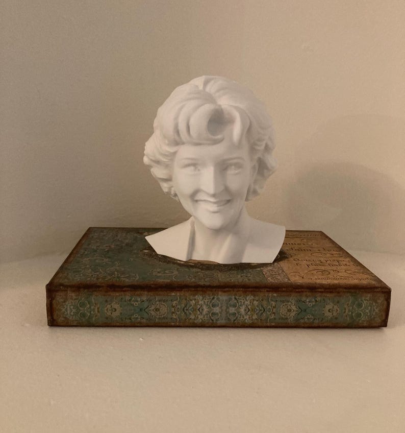 May include: A white bust of a smiling woman with curly hair rests on a rectangular box. The box has a teal and brown patterned design. The bust is a decorative item.