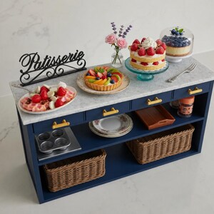 Pu&ograve; includere: Un'isola da cucina in miniatura blu navy con piano in marmo bianco, maniglie dorate e due cesti in vimini. La superficie presenta un'insegna nera Patisserie, un waffle con fragole, una crostata di frutta e diverse torte in miniatura.