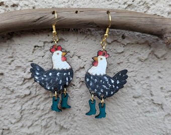 May include: A pair of whimsical earrings featuring hand-painted chickens with white heads, black and white speckled bodies, red combs, and turquoise boots. The earrings hang from gold-colored hooks, showcasing a unique and playful design.