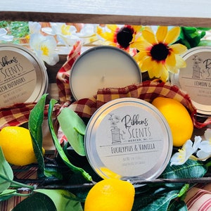 May include: Three silver tin candles with white lids and brown labels. The labels have the text "Ribbons & Scents" and the candle scents are "Fresh Outdoors", "Eucalyptus & Vanilla", and "Lemon Lavender". The candles are surrounded by lemons, white flowers, and a red and white checkered fabric.