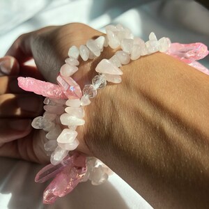 Könnte beinhalten: Zwei Armbänder aus rosa und weißen Kristallperlen. Die Armbänder sind auf dem Handgelenk einer Person übereinander gelegt.